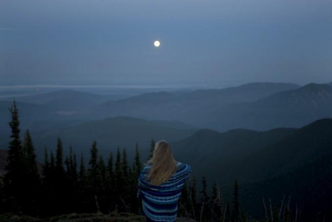 жена умотана у ћебе гледа у планински пејзаж са пуним месецом, поглед отпозади