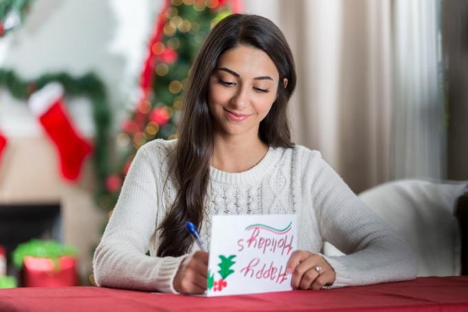 насмејана млада жена седи за столом у својој уређеној дневној соби и концентрисано гледа док пише писмо пријатељу за Божић