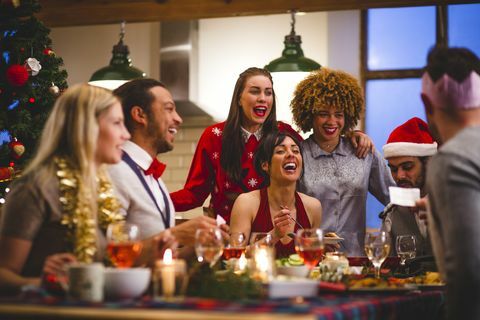 група пријатеља који се смеју док седе за столом и једу и пију за Божић један човек чита виц са божићног крекера носе се шешири и божићни џемпери